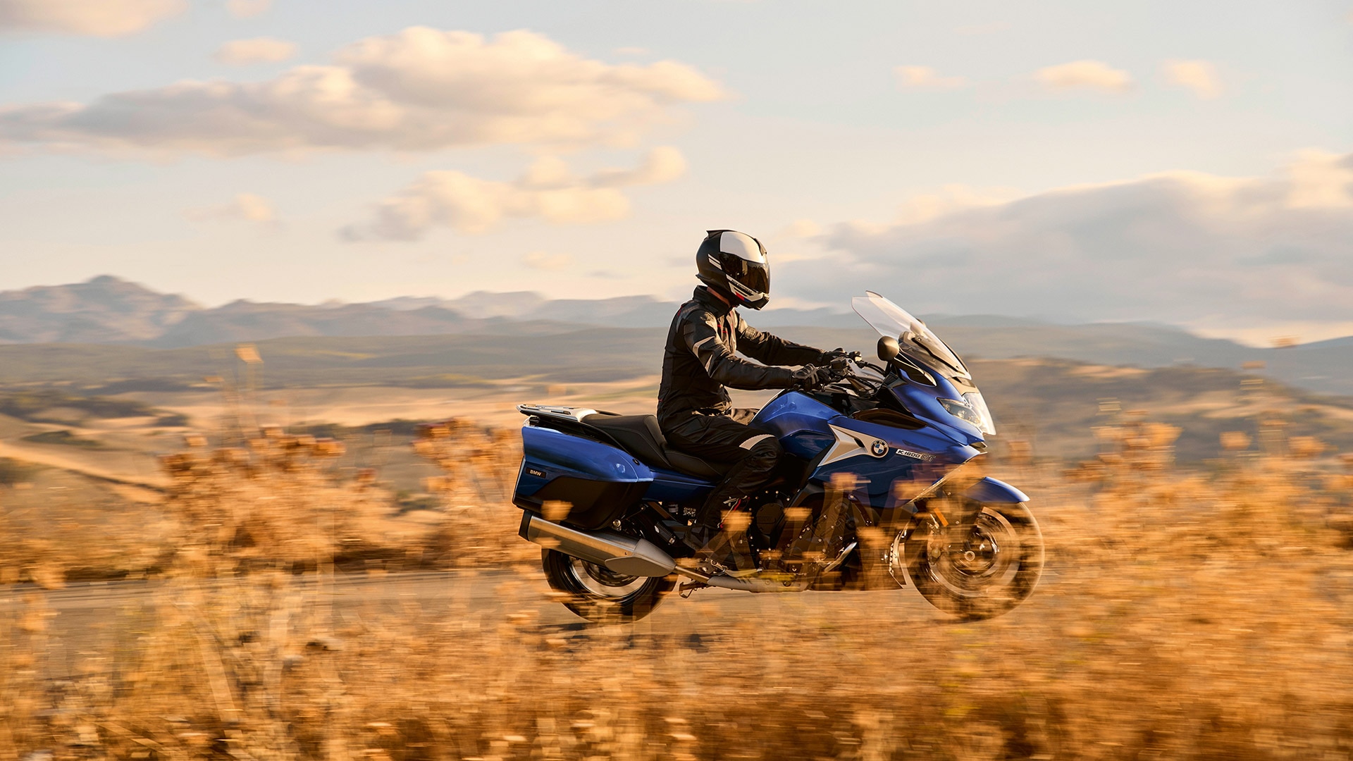 K 1600 GT în Stilul Sport, în mers, în fața unui peisaj deluros uscat, cu iarbă.