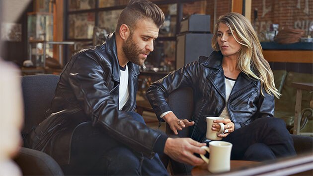 In the image sit a young man in the men’s FlatTwin leather jacket and a young woman in the women's FlatTwin leather jacket across from him.